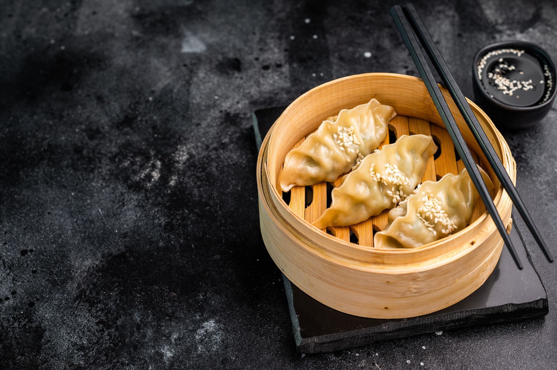 Des raviolis vapeur dans un panier en bambou, accompagnés de baguettes, sur une table sombre, avec de la sauce soja à proximité.