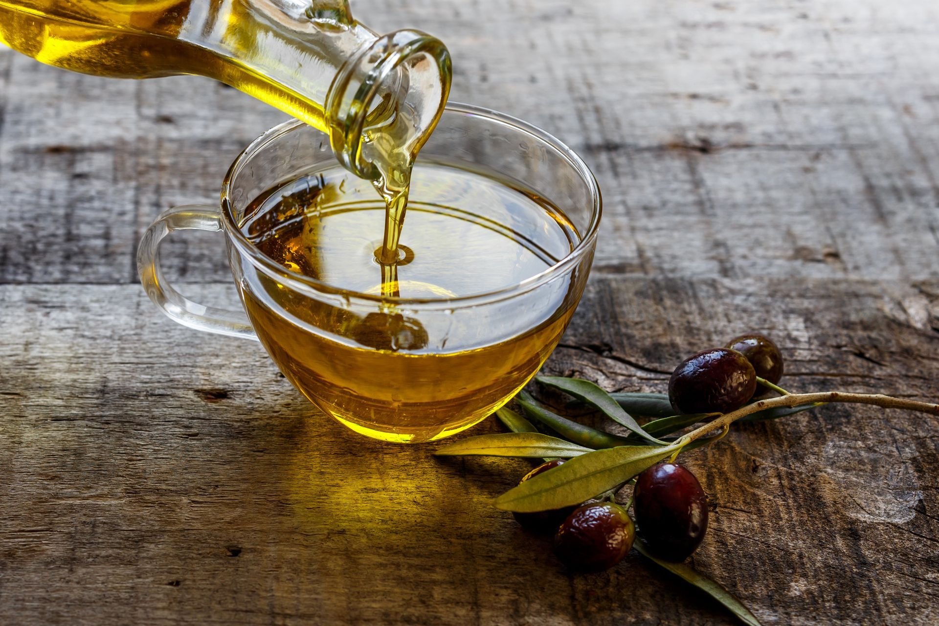 De l'huile d'olive se verse dans un bol en verre posé sur une table rustique en bois, entourée d'olives et de branches d'olivier.