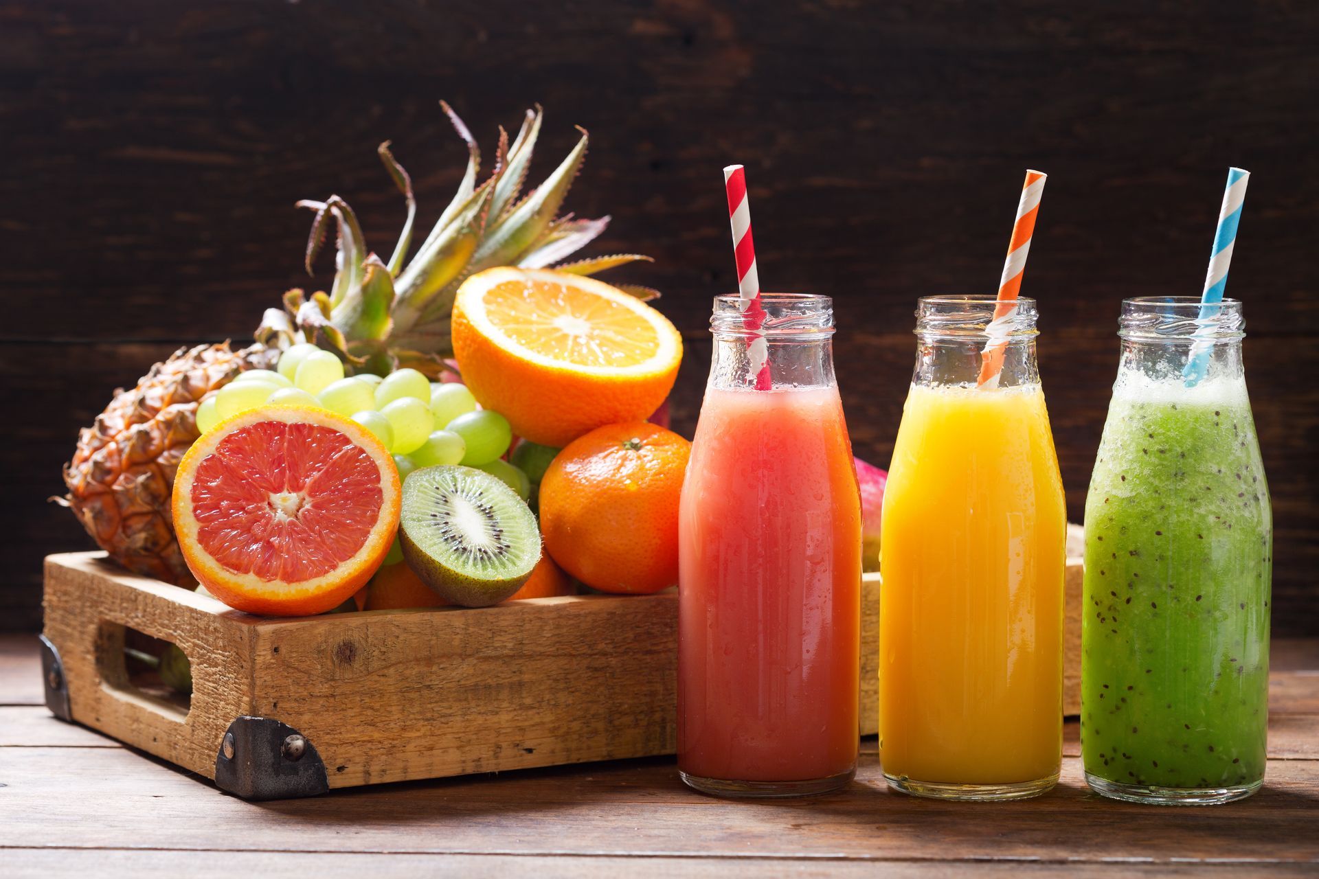 Jus de fruits frais assortis en bouteilles de verre, à côté d'une caisse en bois remplie d'agrumes et d'ananas tranchés.