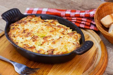 Gratin de fromage cuit au four dans une poêle noire sur une planche en bois, avec du pain et un torchon à carreaux à proximité.