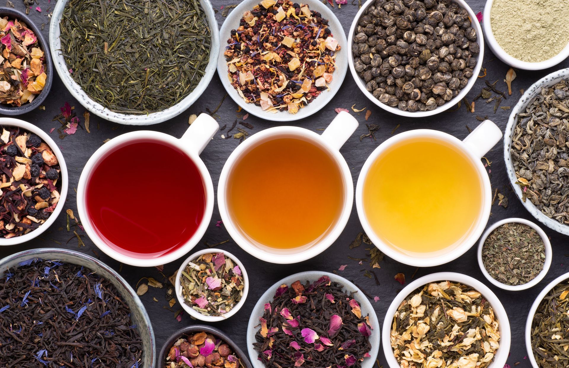 Vue de dessus d'un assortiment de thés en vrac et de trois tasses de thé rouge, ambré et jaune dans des bols.