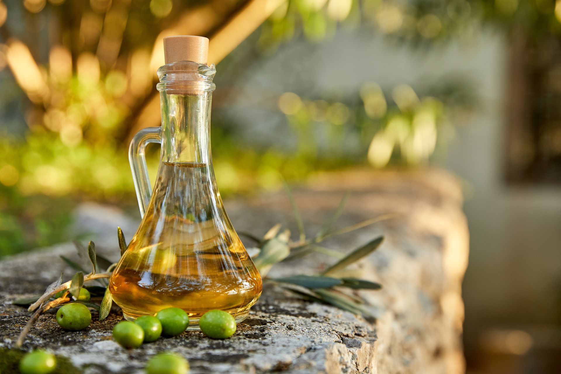 Bouteille en verre d'huile d'olive avec des olives posées sur une surface en pierre à l'extérieur, sous un soleil chaud