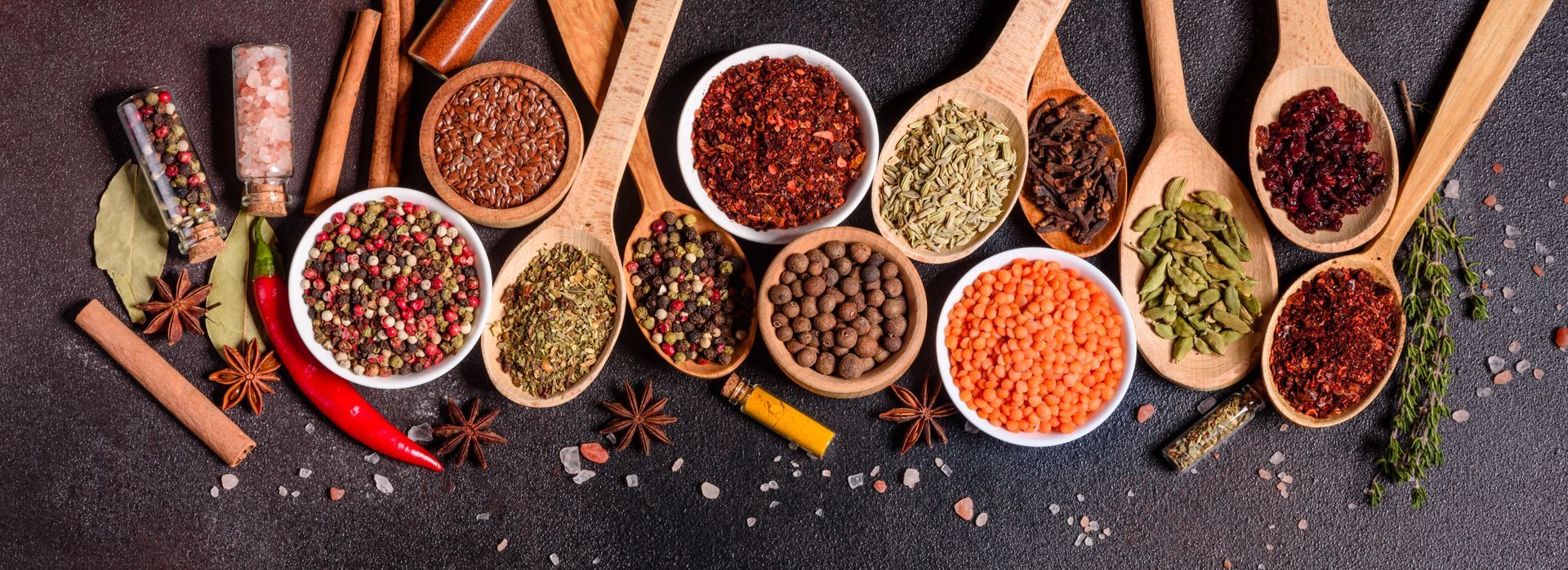 Assortiment d'épices dans des bols et des louches en bois sur une surface sombre, avec des piments rouges et des herbes.
