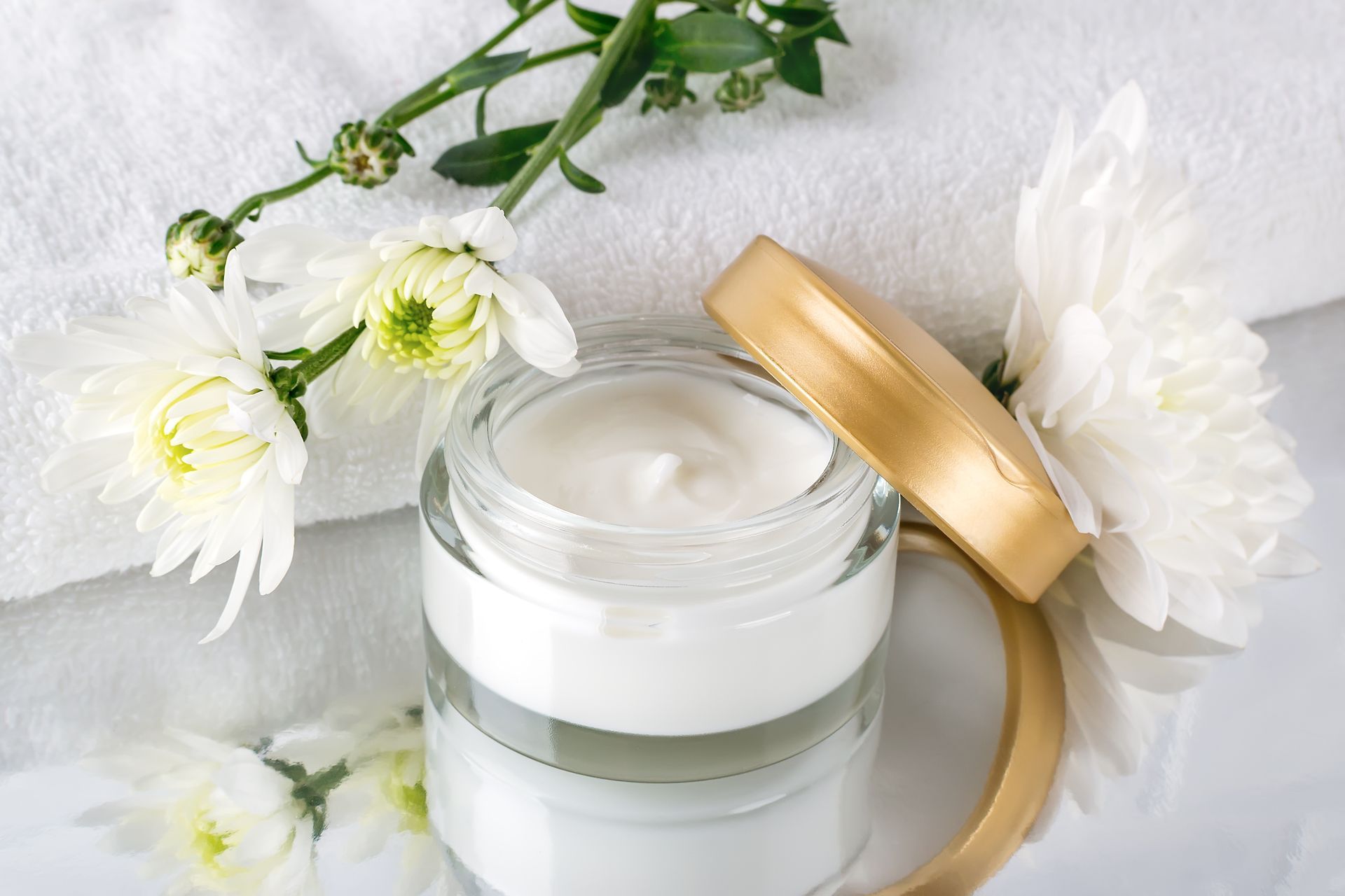 Pot de crème pour le visage blanc à couvercle doré, posé à côté de fleurs blanches sur un fond doux.