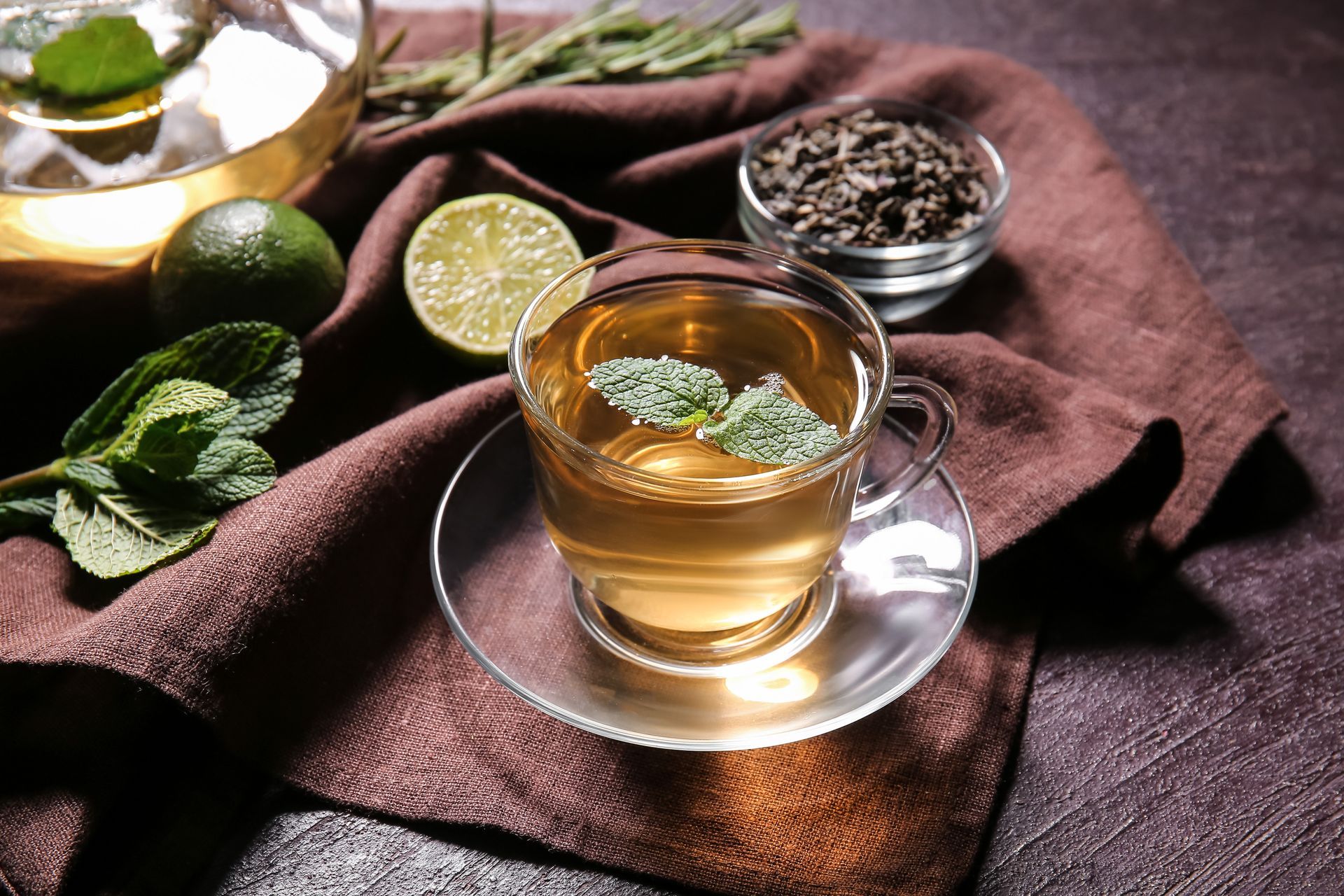 Tisane dans une tasse transparente posée sur un linge brun, avec de la menthe, des citrons verts et du thé en vrac à proximité.