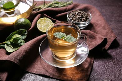 Tasse en verre de tisane à la menthe posée sur un linge brun, à côté de tranches de citron vert et de feuilles détachées.