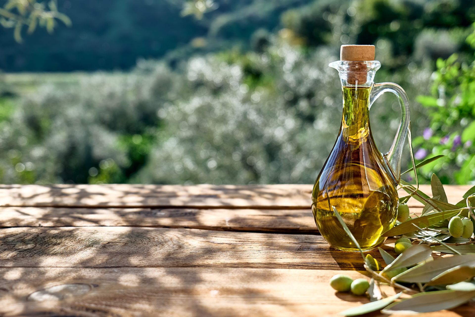 Bouteille d'huile d'olive avec bouchon de liège sur une table en bois ensoleillée, avec des olives et de la verdure floue en arrière-plan.