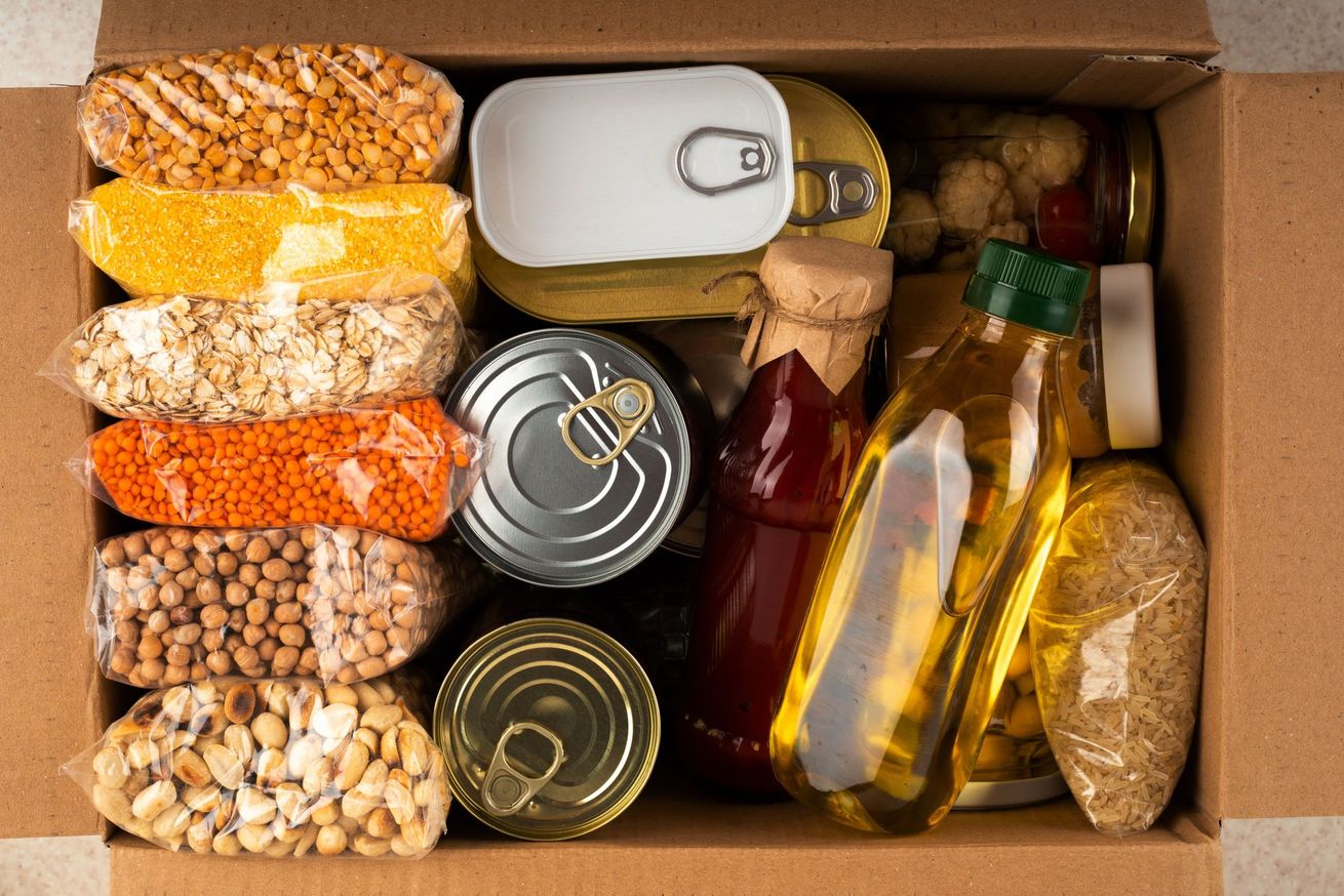 Boîte remplie de produits d'épicerie assortis, comprenant des céréales, des conserves, des pâtes, de l'huile et du pain.
