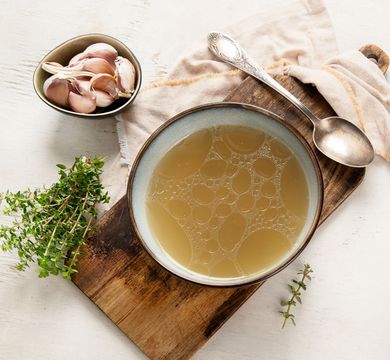 Bol de bouillon clair sur une planche en bois avec de l'ail, des herbes et des cuillères sur un fond de tissu léger