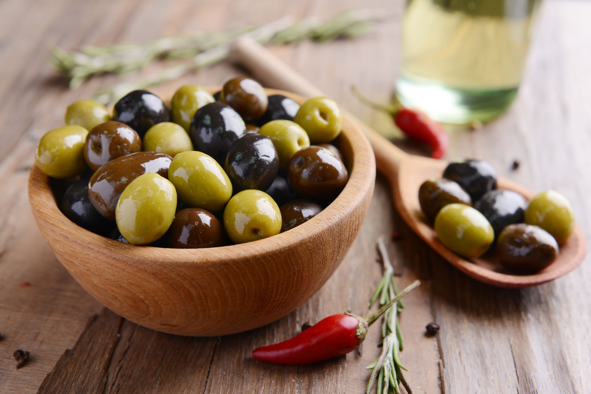 Bol d'olives vertes et noires mélangées sur une table en bois avec des piments rouges et des herbes.