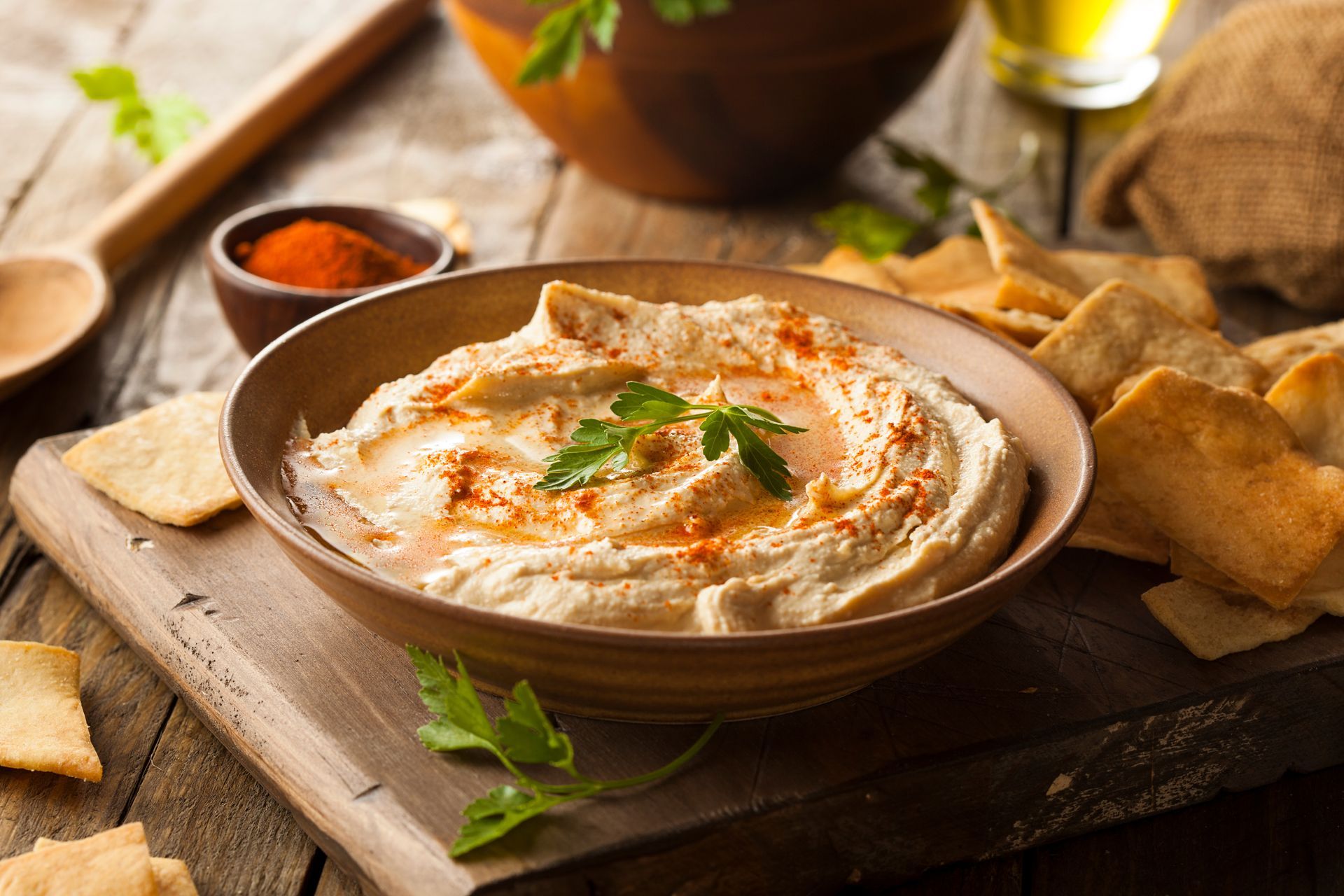 Houmous crémeux servi dans un bol avec des chips de pita sur une planche en bois, garni d'herbes et de paprika