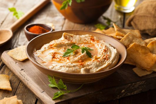 Houmous servi dans un bol avec du paprika et du persil, accompagné de chips de pita sur une planche en bois.