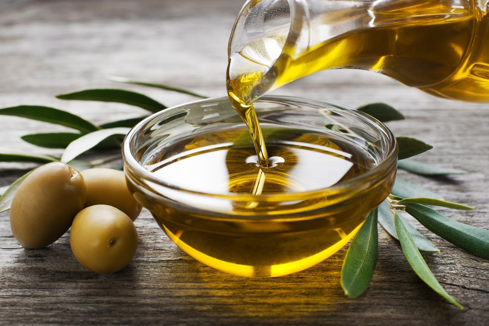 De l'huile d'olive est versée dans un bol en verre contenant des olives et des branches d'olivier, sur une table rustique.