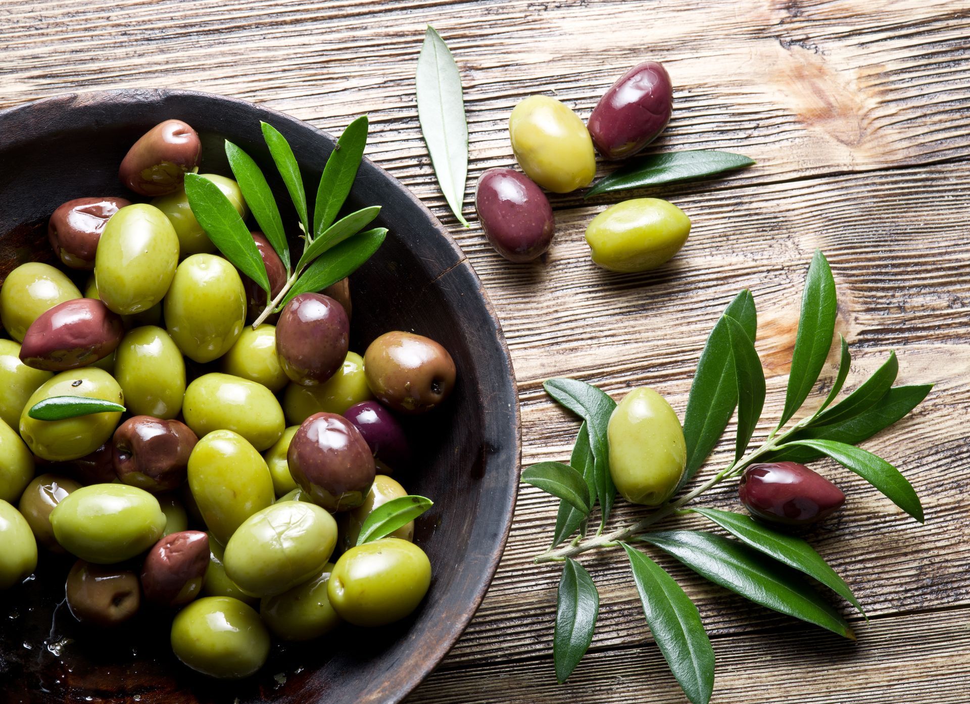 Olives vertes et violettes assorties dans un bol noir posé sur une surface en bois rustique, avec des brins d'olivier à proximité.