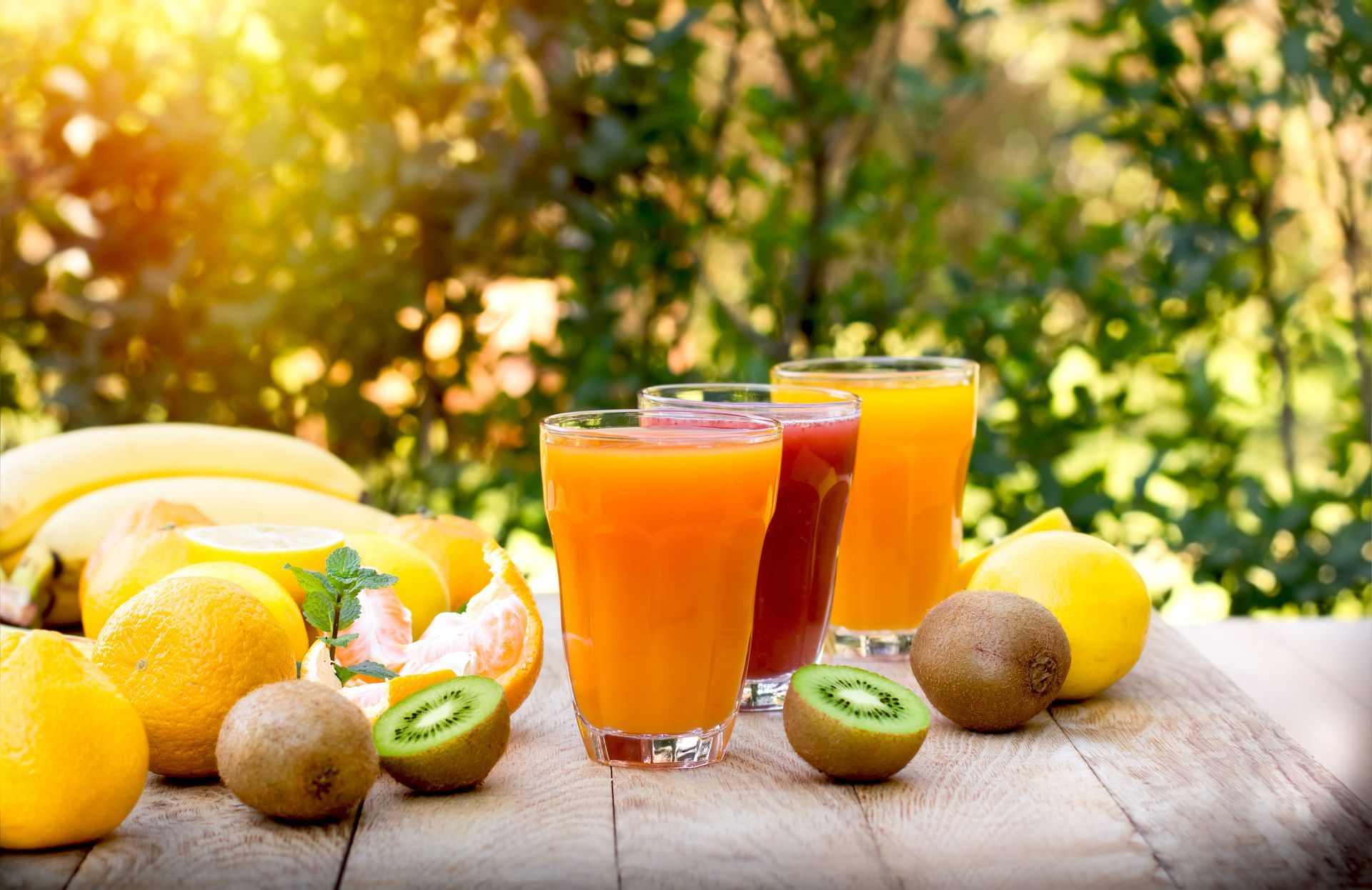 Smoothies aux fruits frais servis dans des verres sur une table en bois, avec des oranges, des bananes, des kiwis et des citrons, à l'extérieur.