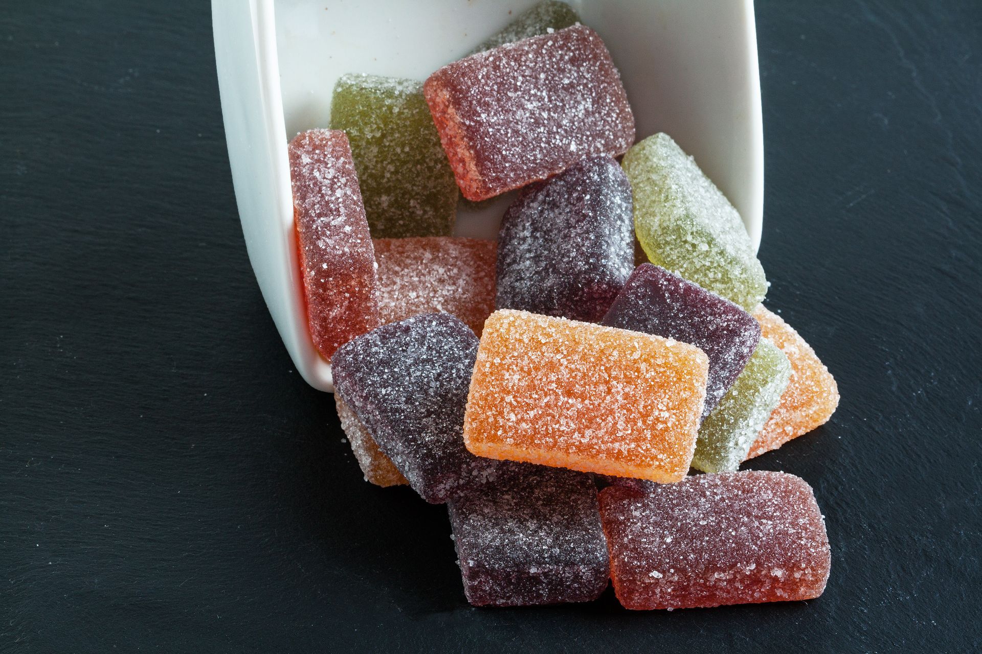 Des bonbons gélifiés colorés enrobés de sucre se répandent d'une tasse blanche renversée sur une surface sombre.