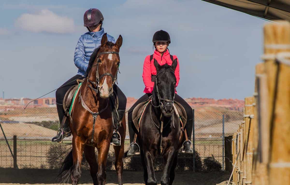 Dos personas montando a caballo al aire libre, un caballo marrón y otro negro.