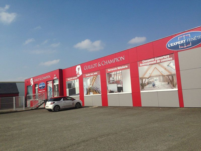 Façade rouge avec de grandes vitrines et une voiture argentée garée sous un ciel bleu