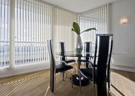 Salle à manger moderne avec table en verre, chaises noires, vase en centre de table et stores verticaux laissant entrer la lumière du jour.