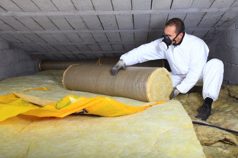 Un ouvrier en combinaison de protection blanche inspecte une couverture isolante jaune enroulée dans un vide sanitaire de grenier.