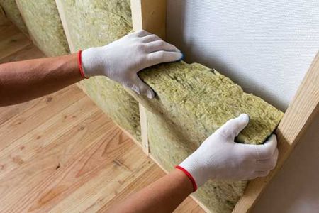 Des mains gantées installent des rouleaux d'isolant jaunes dans une cavité murale sur un plancher en bois.