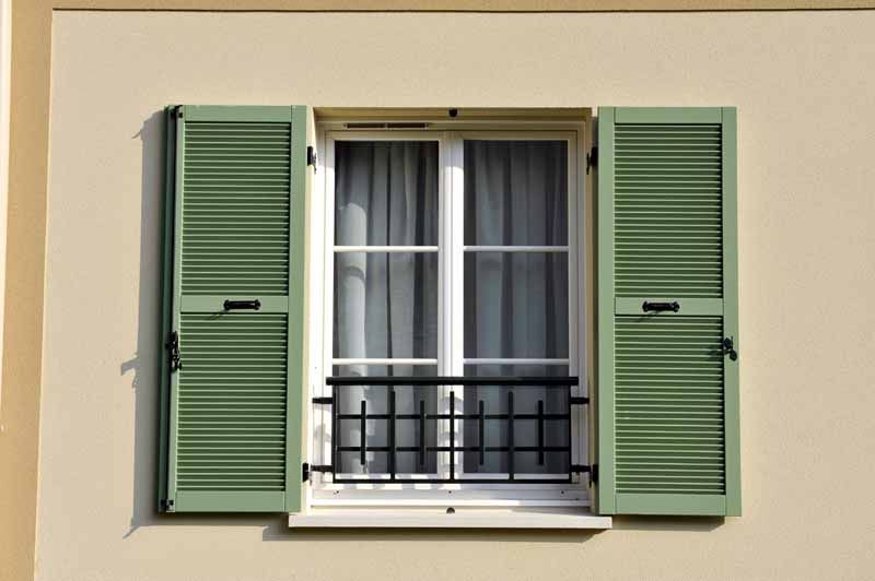 Volets verts encadrant une fenêtre à cadre blanc avec rambarde de balcon noire sur un mur beige