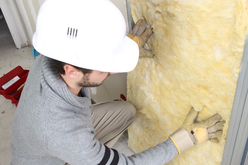 Ouvrier installant de l'isolant en fibre de verre jaune dans une cavité murale, portant un casque et des gants de chantier.