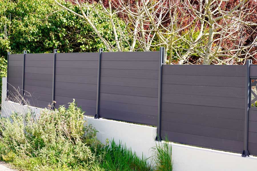 Clôture d'intimité gris foncé le long d'un mur de soutènement blanc, avec de la verdure et des arbres en arrière-plan.