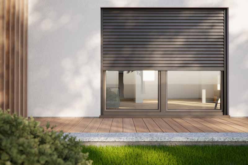 Extérieur de maison moderne avec un grand volet roulant gris au-dessus de portes coulissantes en verre et une terrasse en bois.