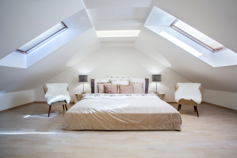 Chambre mansardée lumineuse avec un lit bas, deux chaises blanches et des fenêtres de toit des deux côtés