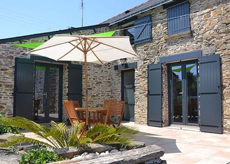 Terrasse avec table et chaises en bois sous un parasol blanc, à côté d'une maison en pierre aux portes-fenêtres ouvertes.