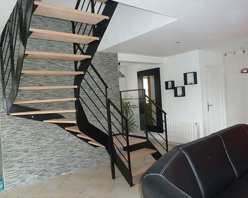 Salon moderne avec escalier ouvert, rampe en métal noir et canapé foncé sous un plafond mansardé