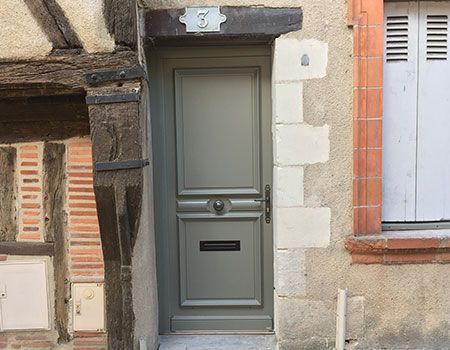 Porte d'entrée grise encastrée dans une étroite façade en pierre, avec des volets orange et une petite fente à lettres.