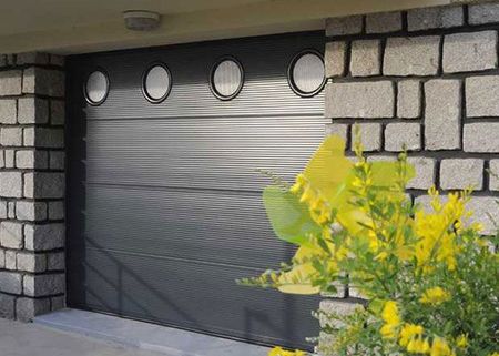 Bâtiment en pierre avec une porte de garage sectionnelle gris foncé et des fleurs jaunes au premier plan