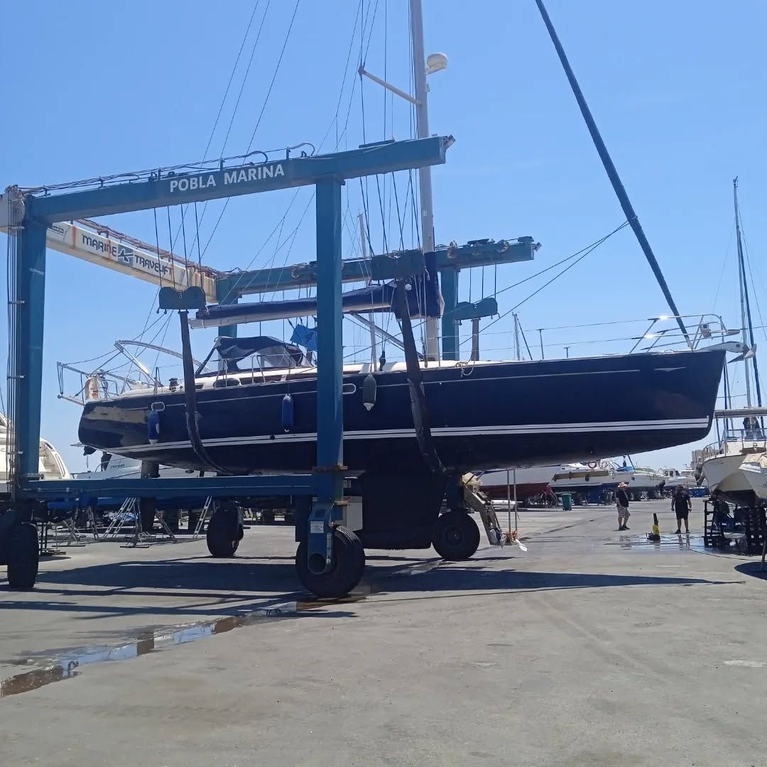 Velero azul oscuro izado por una grúa en un puerto deportivo. Cielo azul sobre sus cabezas.