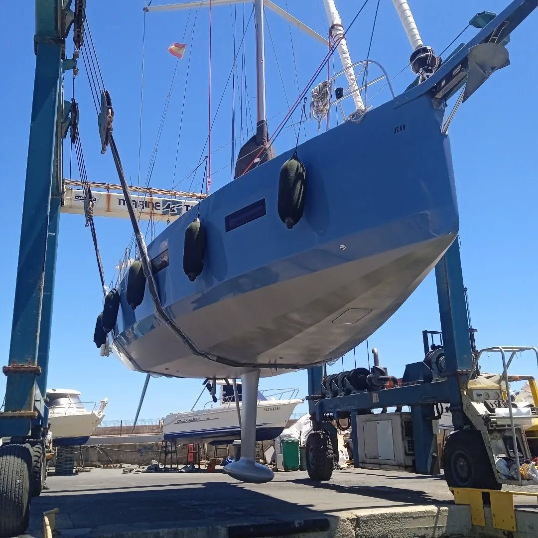 Un velero izado por una grúa en un astillero. El casco es azul y gris, con el cielo azul como fondo.