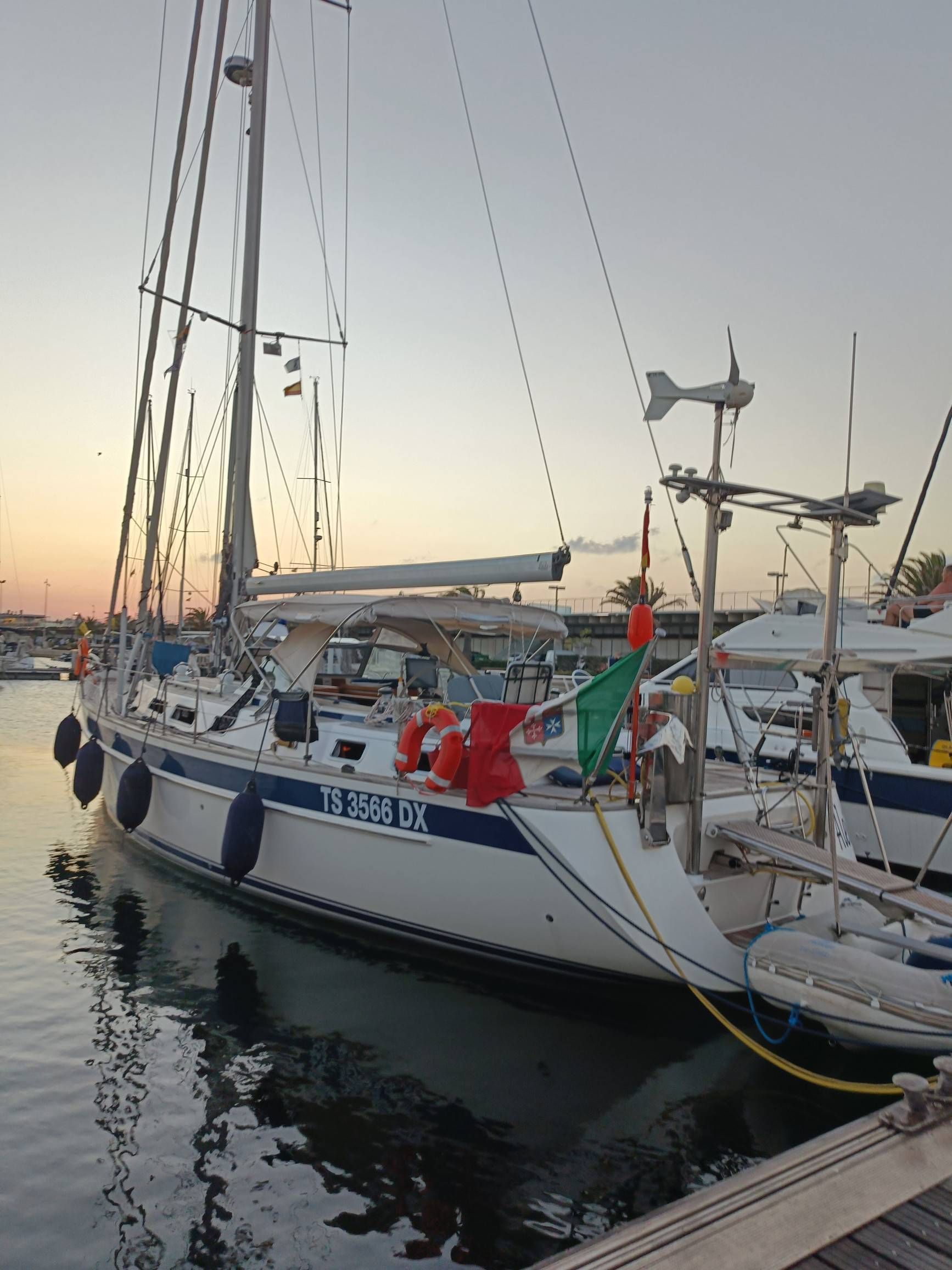 Velero atracado al anochecer, casco blanco, franja azul, bandera italiana, indicador de viento.
