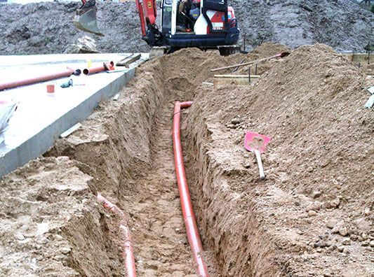 In einem Graben im Boden wird ein rotes Rohr verlegt.