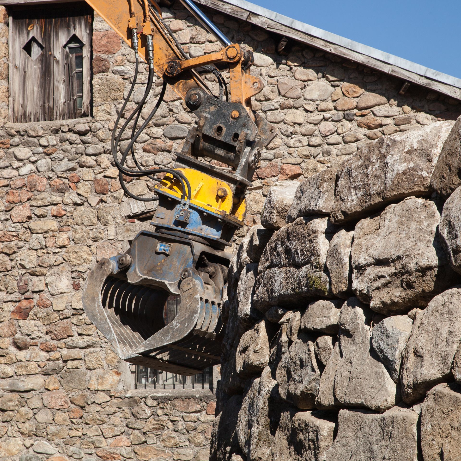 Une pince de démolition montée sur une excavatrice abat un mur de pierre à côté d’un vieux bâtiment