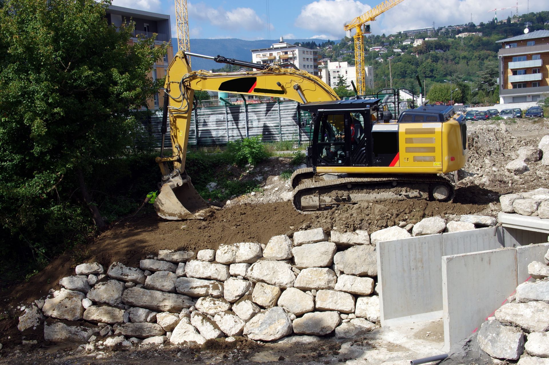 Une pelleteuse jaune creuse sur un chantier, à côté d’un mur de soutènement en pierre