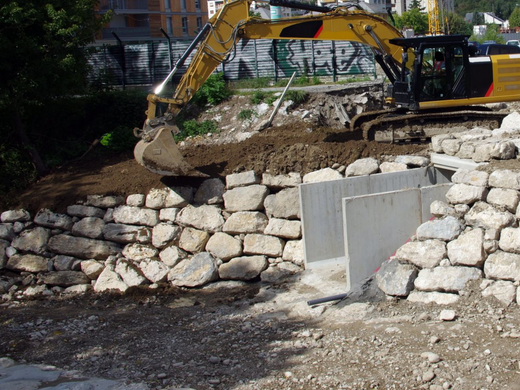 Une excavatrice jaune creuse derrière un mur de soutènement en pierre sur un chantier