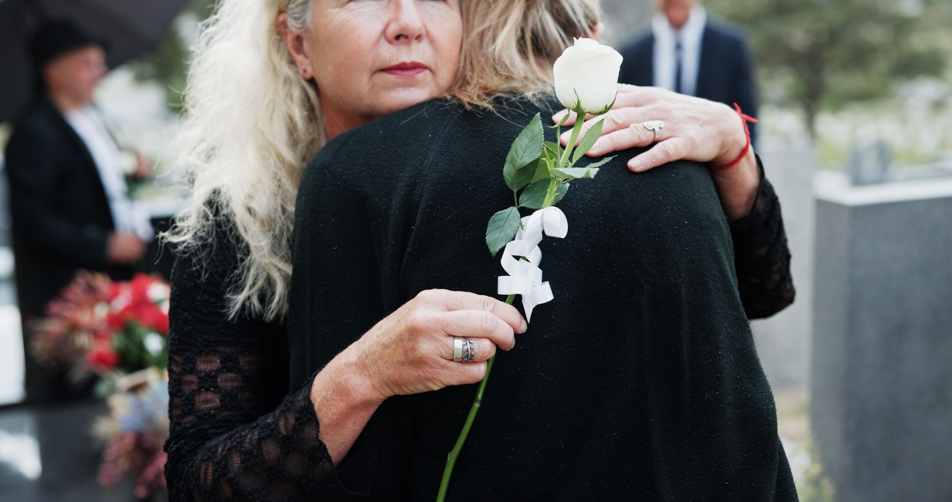 Personne vêtue de noir tenant une rose blanche lors d’une cérémonie commémorative et enlaçant une autre personne