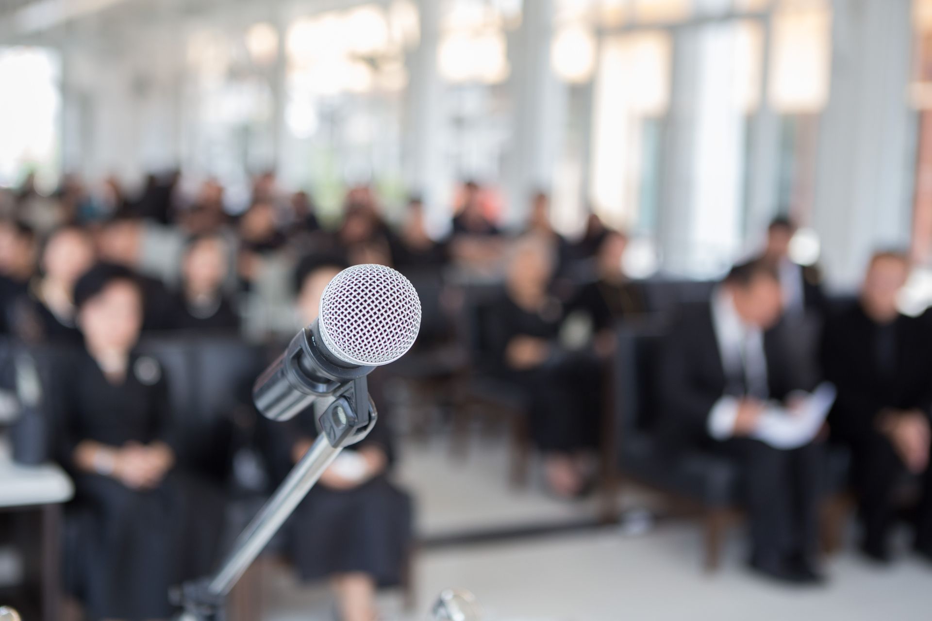 Microphone sur pied au premier plan, avec un public assis flou dans une salle lors d'une cérémonie funéraire