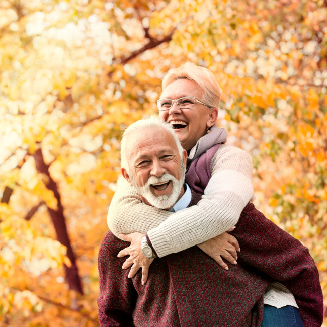 Un couple âgé souriant s’enlace en plein air en automne, avec des feuilles dorées en arrière-plan