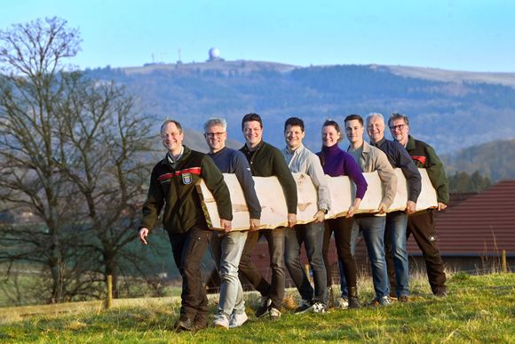 Acht Personen stehen lächelnd im Freien auf einem Hügel und tragen gemeinsam ein langes, helles Holzbrett.