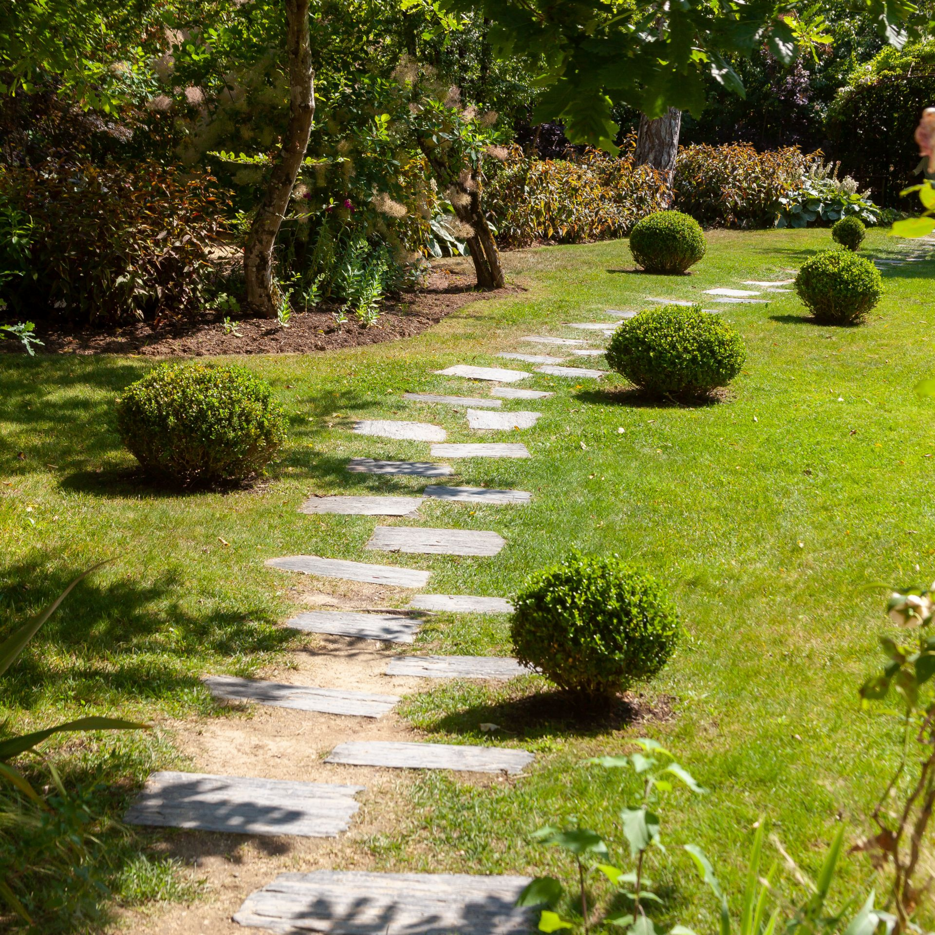 Un chemin de pierre serpente dans un jardin verdoyant, entouré de buissons et d'arbres.
