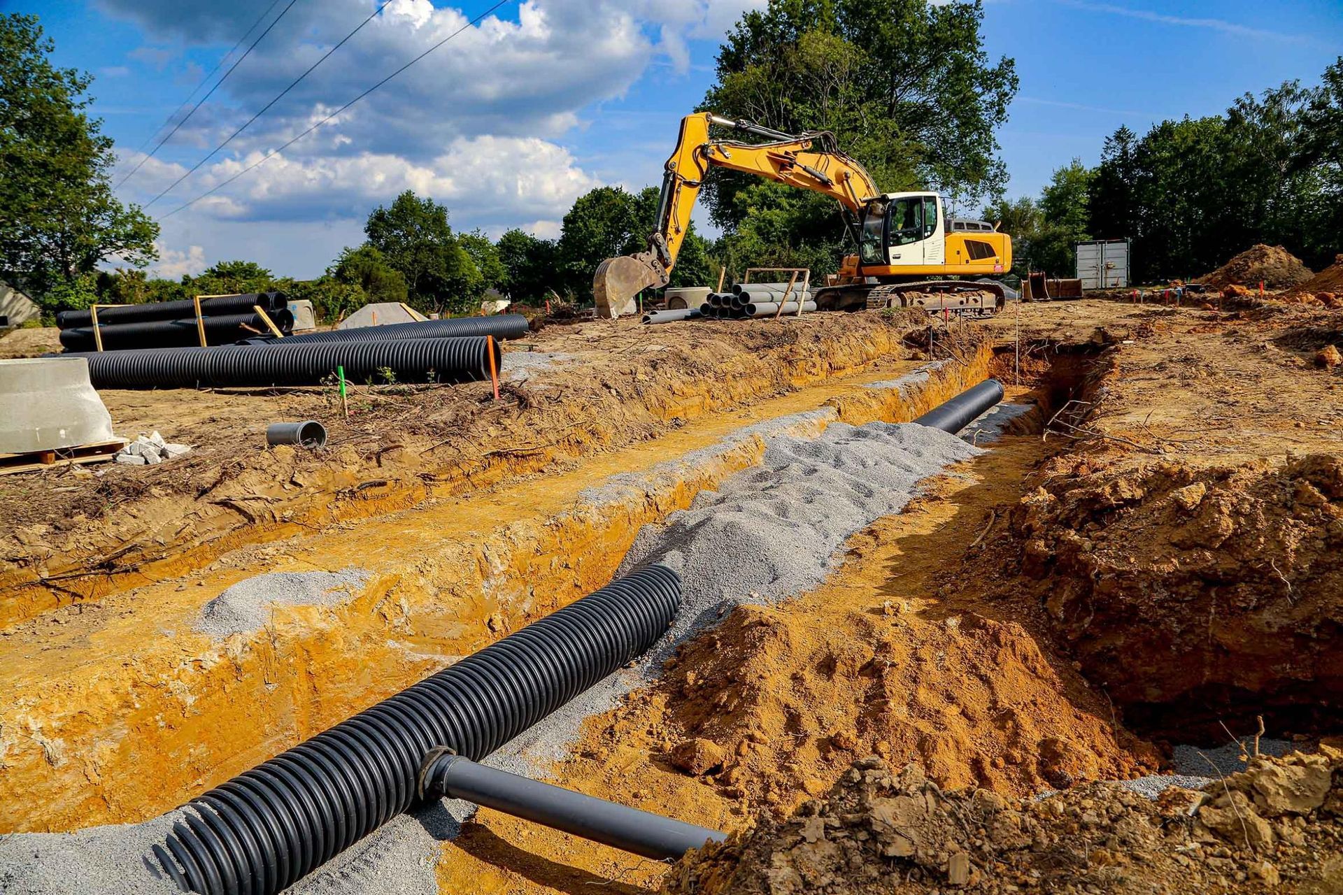 Une excavatrice installe de grosses canalisations de drainage sur un chantier boueux, sous un ciel bleu