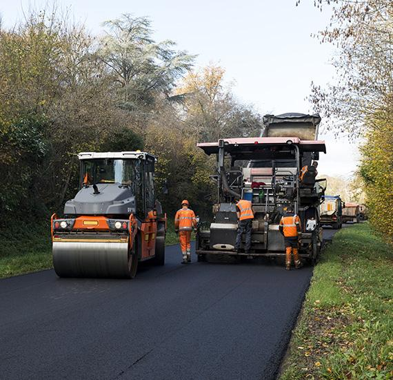 Des ouvriers routiers qui goudronnent une rue avec deux finisseurs d’asphalte, sur une route bordée d’arbres