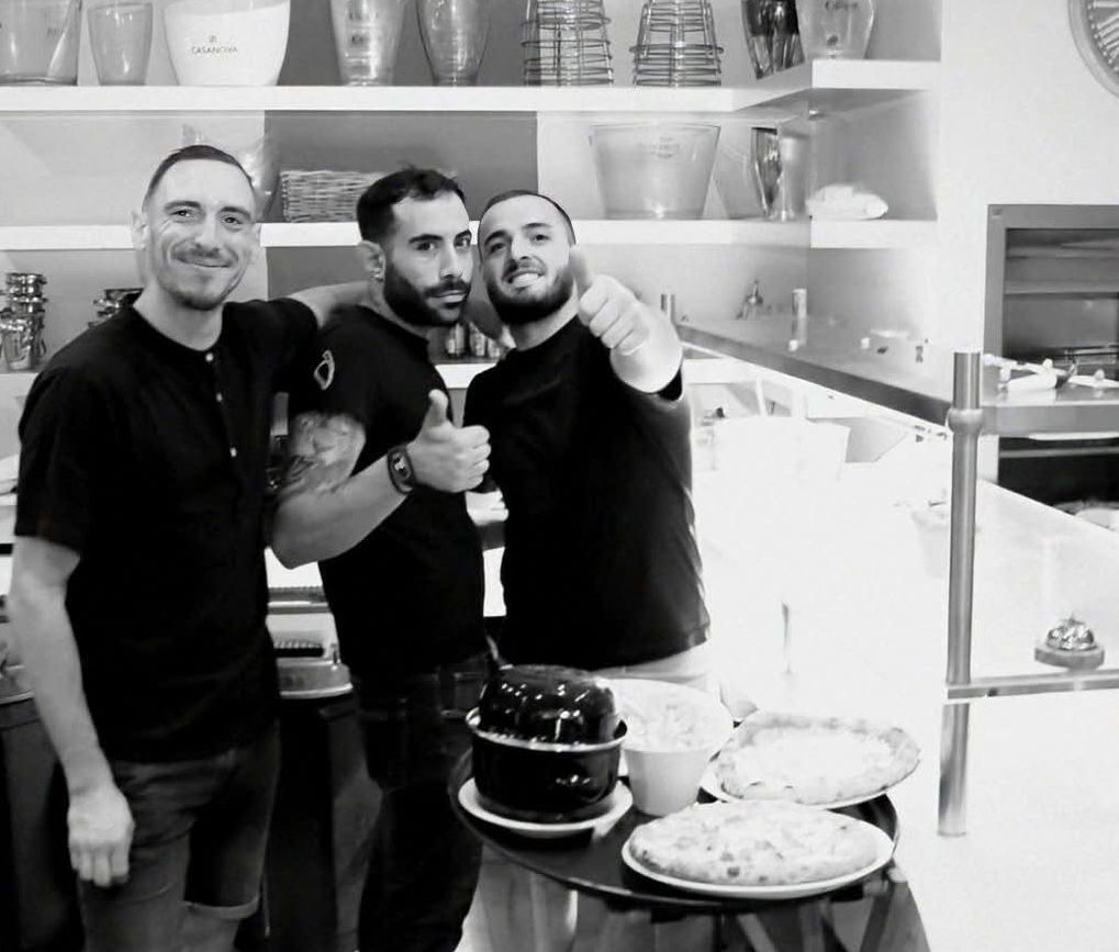 Trois personnes posent dans une boulangerie ou un café lumineux, souriant derrière un comptoir garni de gâteaux et d'étagères remplies de vaisselle.