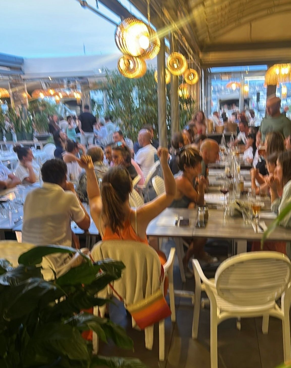 Café en terrasse animé au crépuscule, les clients étant attablés à de longues tables sous des guirlandes lumineuses chaleureuses.